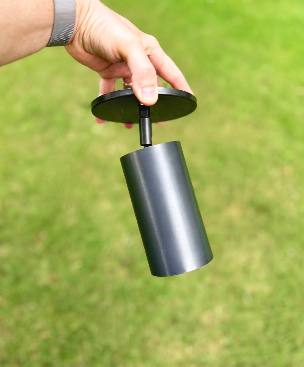 A hand holds the Long Made Co. Highland Adjustable Spot, a minimalist brass spotlight with a cylindrical metallic body and round black top, set against blurred green grass.