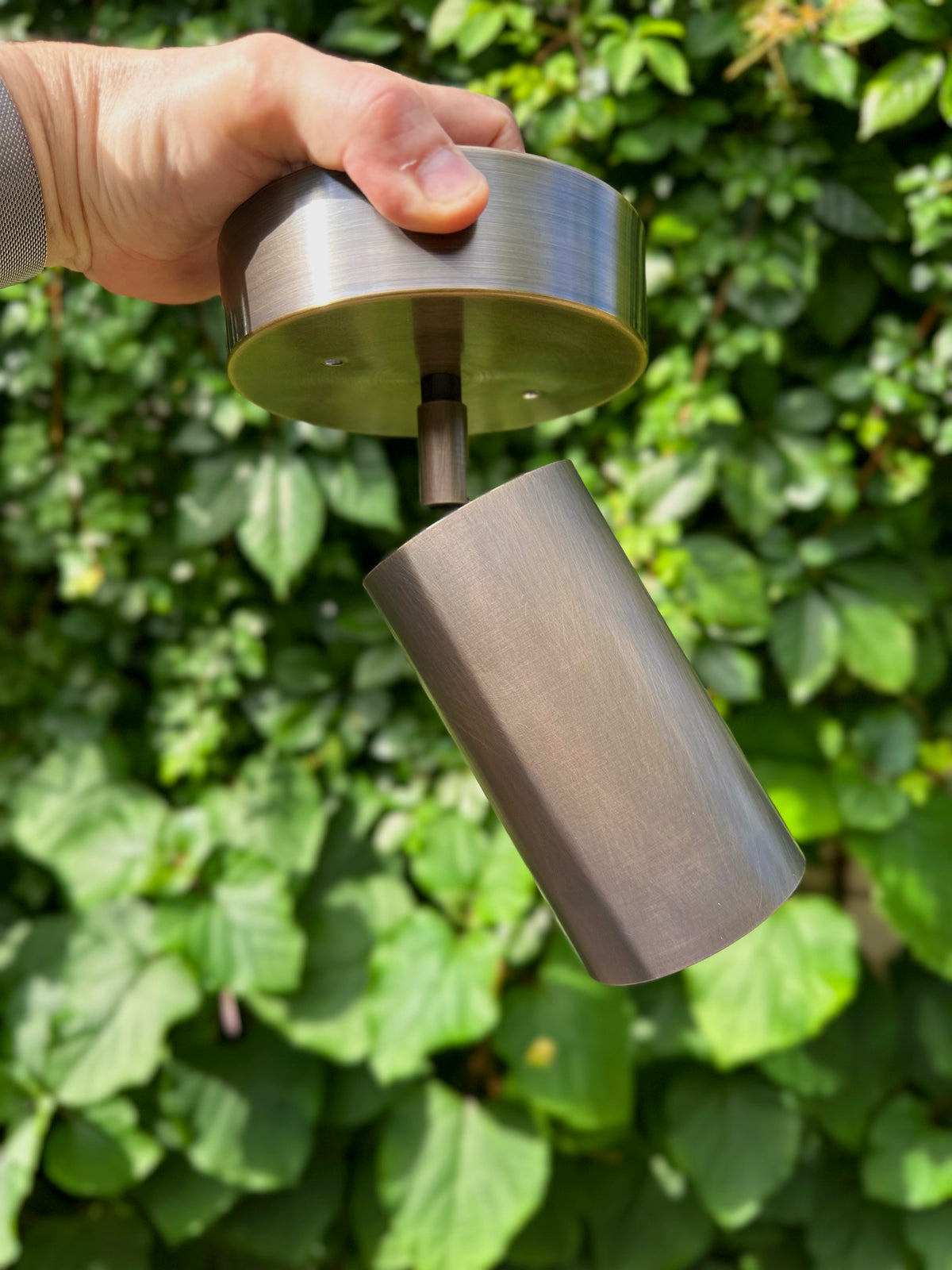 A hand holds the Long Made Co. Highland Adjustable Chunky Spot with a metal finish, shown against a leafy green background—an elegant choice for minimalist lighting.