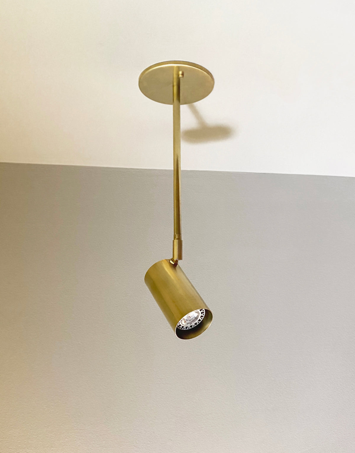The Brass Cylinder Drop Spot - Side Mount by Long Made Co. features a brass cylinder hanging from a round base, providing directional ceiling illumination—ideal for mid-century modern spaces with white and gray accents.