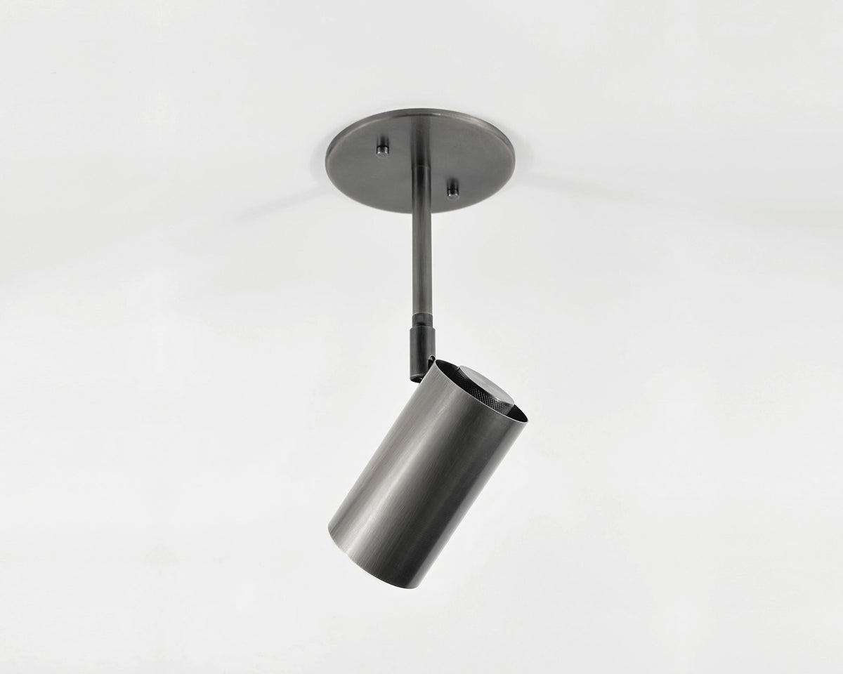 The Brass Cylinder Drop Spot - Side Mount by Long Made Co. is a modern cylindrical metal ceiling spotlight with a black finish, attached to a round base by a straight rod and set against a plain white background.