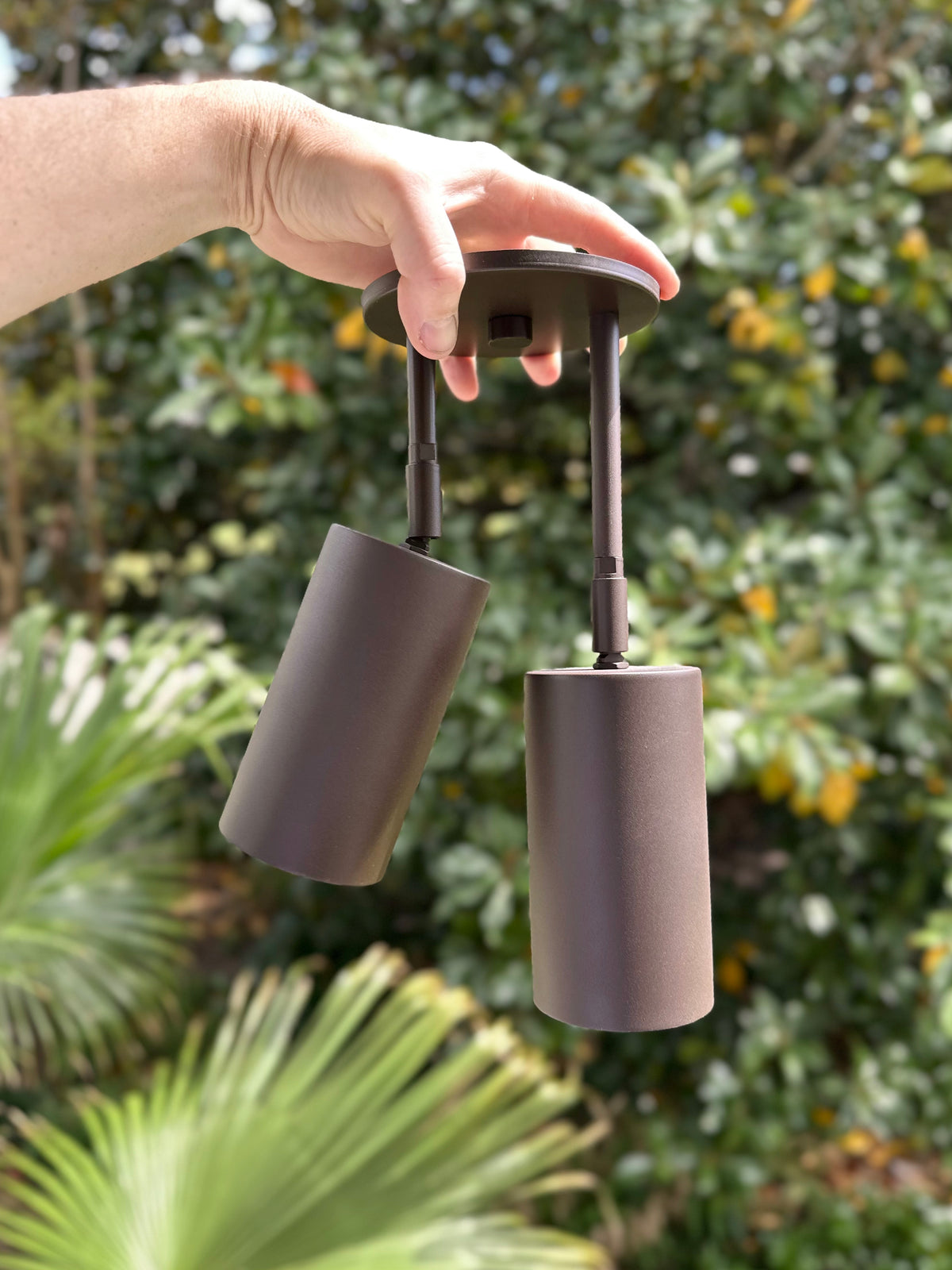 A hand holds the Cove Double Drop Spot, a modern outdoor light fixture from Long Made Co., against a blurred backdrop of green foliage and trees.