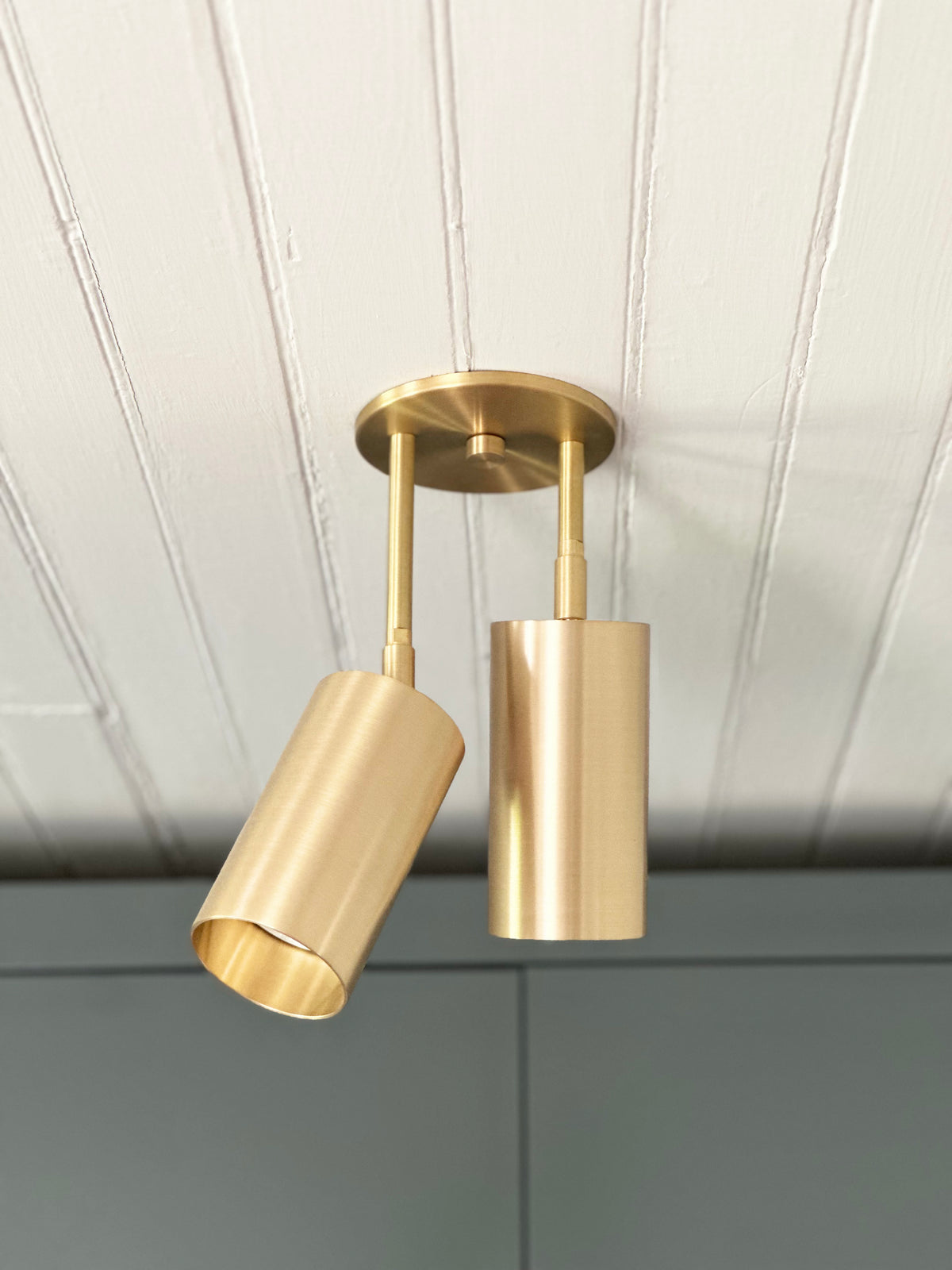 The Highland Double Drop Spot by Long Made Co. is a brass double cylinder ceiling fixture with two adjustable lights, mounted on a white paneled ceiling above a gray wall or cabinet.
