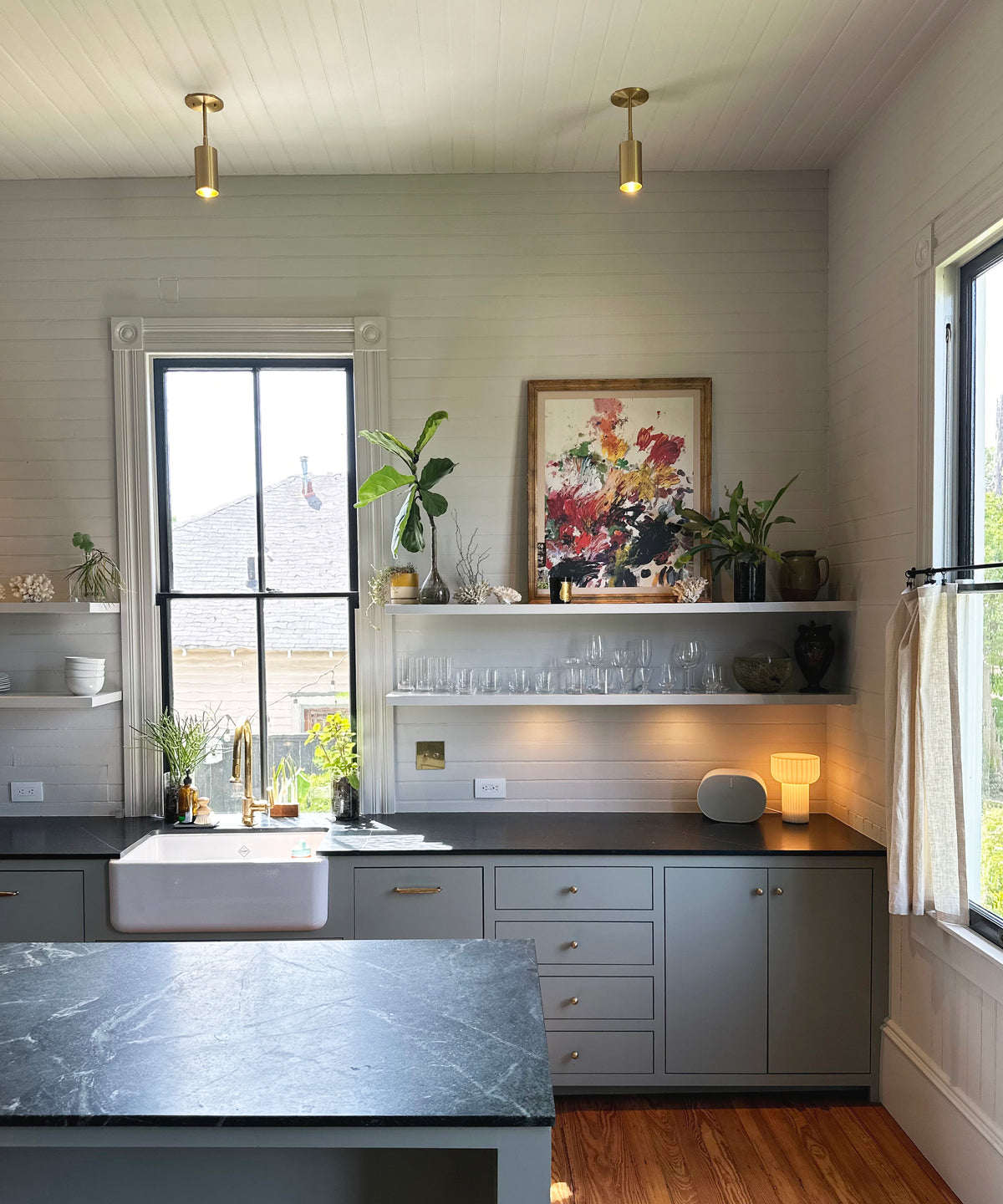 Bright kitchen with pale gray cabinets, marble island, farmhouse sink, open shelves, art, and small lamp—illuminated by Long Made Co.’s Highland Drop Spot brass spotlights and sunlight streaming through the window.