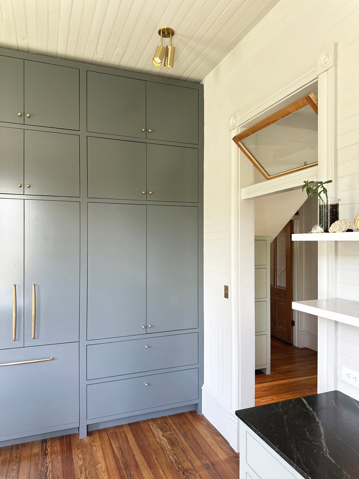Modern kitchen with tall gray cabinets, brass hardware, wood floors, white walls, and a black countertop. A shelf holds decor and a plant. Gold Highland Double Drop Spot lights by Long Made Co. add style to the space.