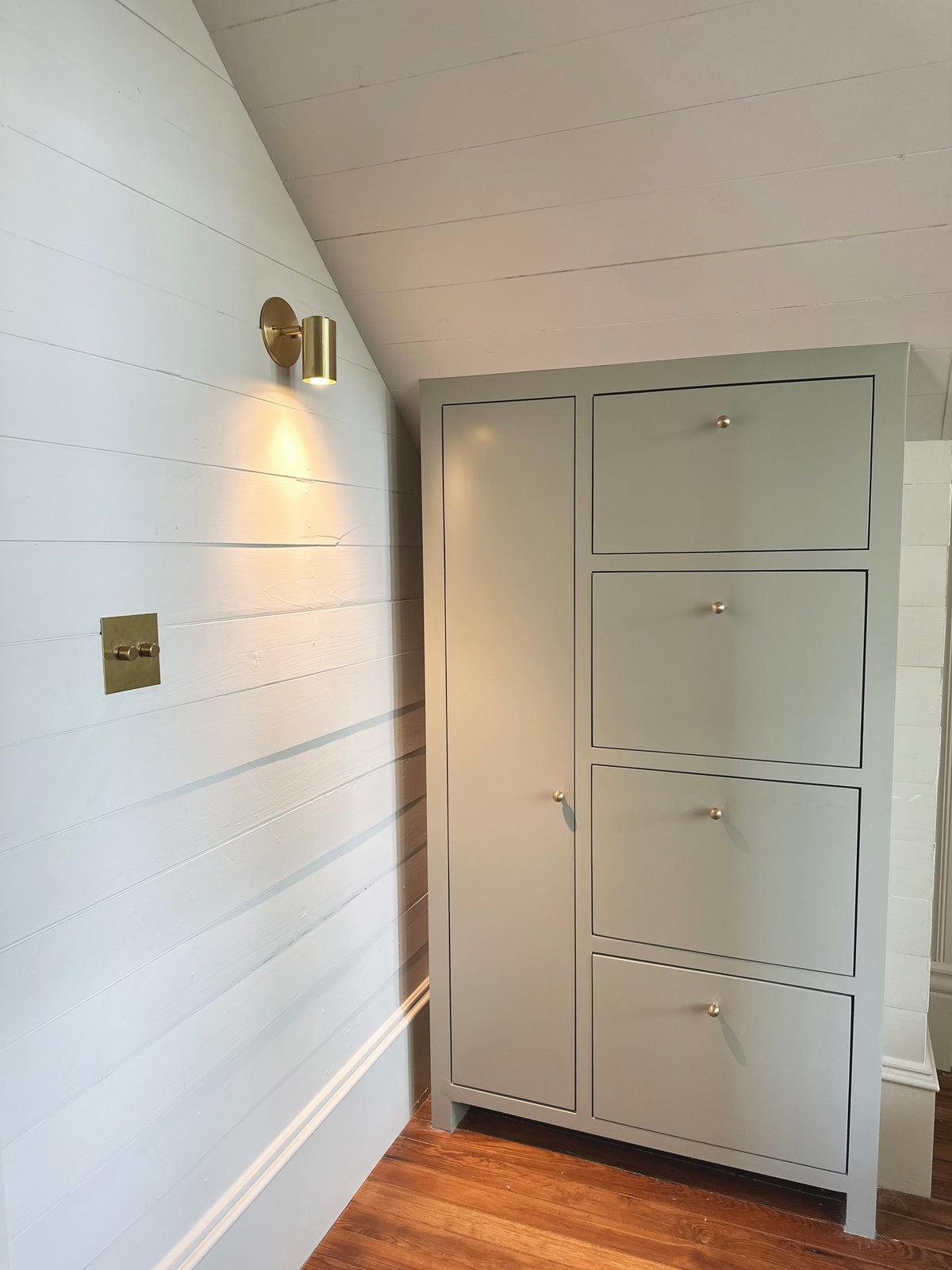 A light green cabinet with four drawers and a tall door stands in a room with white shiplap walls, wood floor, and warm light from the Brass Cylinder Wall Lamp by Long Made Co., creating an inviting atmosphere.