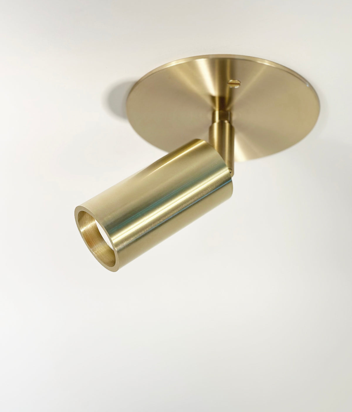The Micro Adjustable Spot by Long Made Co. is a modern, cylindrical brass ceiling spotlight with an ultra-thick inset head, mounted on a round brass base and angled downward for sleek, minimalist appeal.