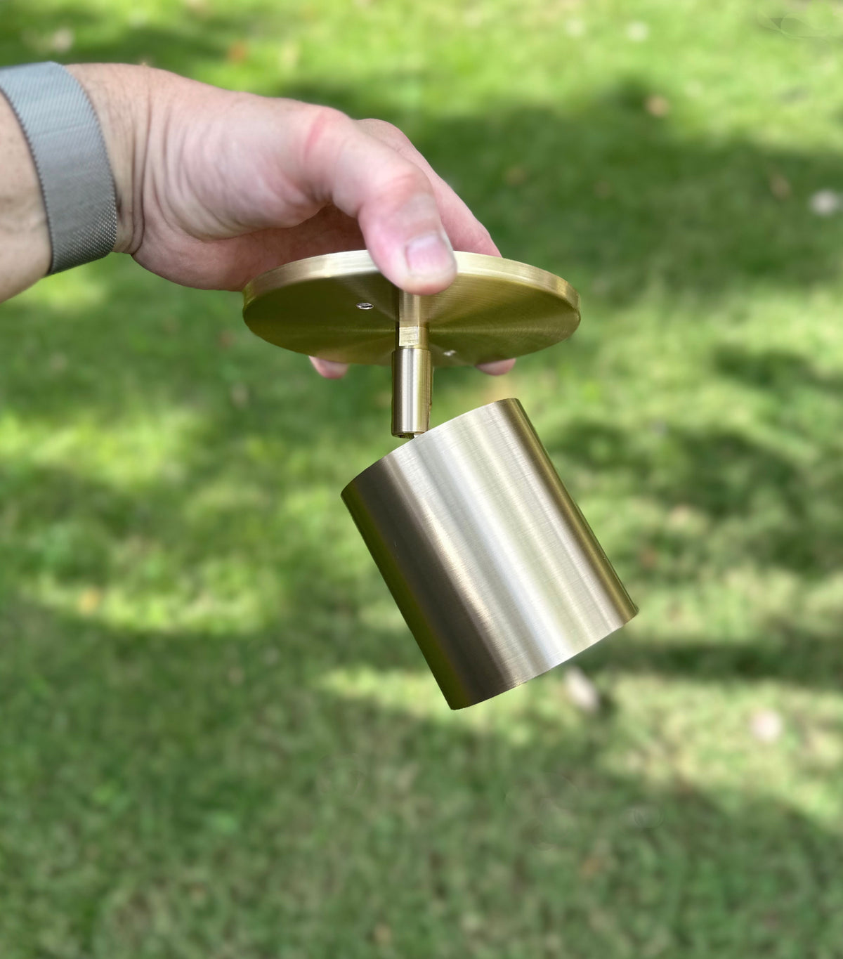 A hand holds the Highland Adjustable Spot - Stocky by Long Made Co. outdoors, with grass in the background. The modern brass fixture has a minimalist cylindrical shade and a round base.