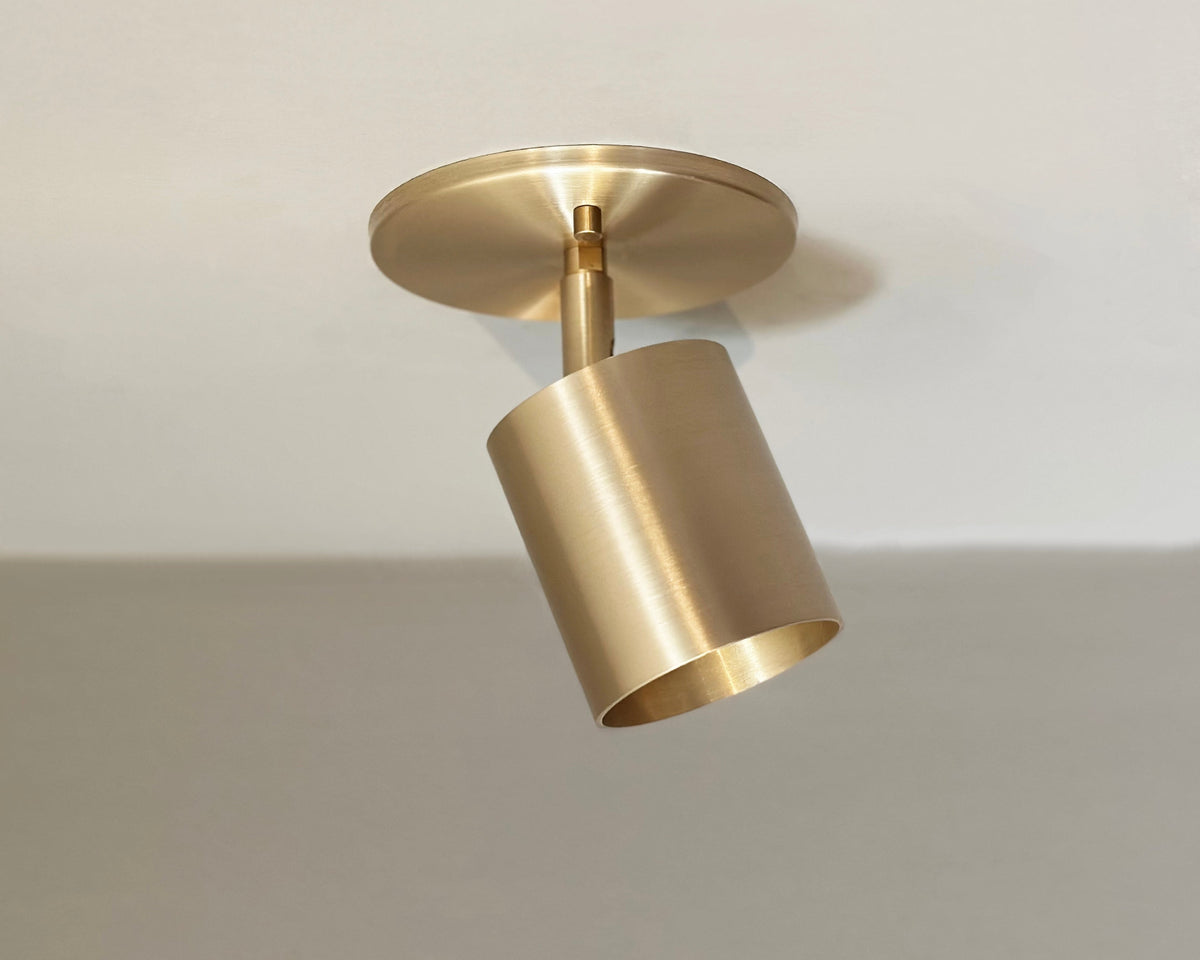 The Highland Adjustable Spot - Stocky by Long Made Co. is a brass ceiling spotlight with a round base and cylindrical shade, angled downward. Its minimalist adjustable design suits modern white ceilings perfectly.
