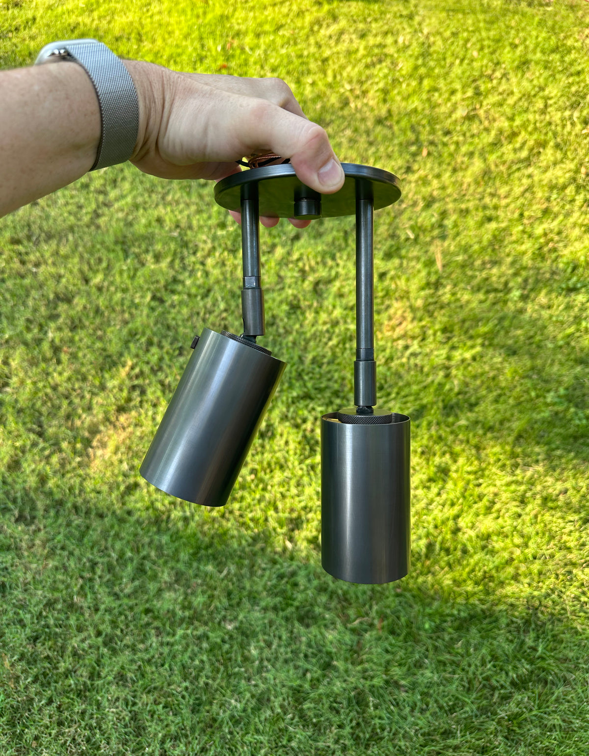 A hand holds the Long Made Co. Brass Cylinder Double Drop Spot, a modern black outdoor fixture with two cylindrical lights, set against green grass.