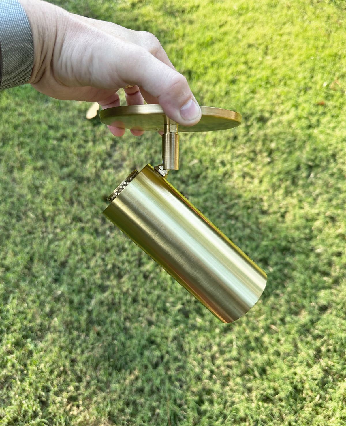 A hand holds the Long Made Co. Brass Cylinder Adjustable Spot - Side Mount w/ Long Shade, featuring a circular mounting plate, against green grass.