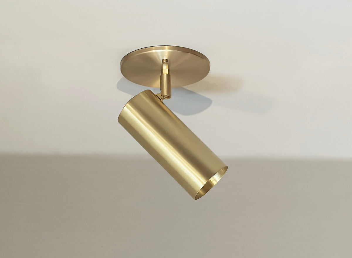 The Brass Cylinder Adjustable Spot - Side Mount w/ Long Shade by Long Made Co. features a modern cylindrical brass design on a round base, angled downward for versatile mid-century modern lighting against any light-colored ceiling.