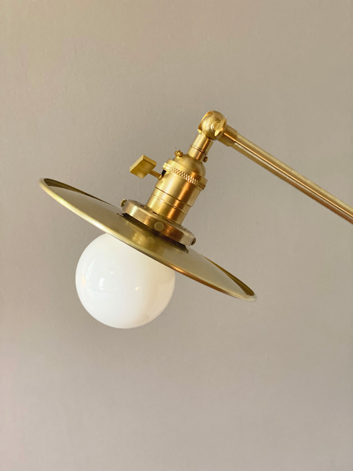 A detailed view of the Task Articulating Chandelier by Long Made Co., featuring solid brass construction, a round white bulb, and a flat circular shade set against a plain light gray background.