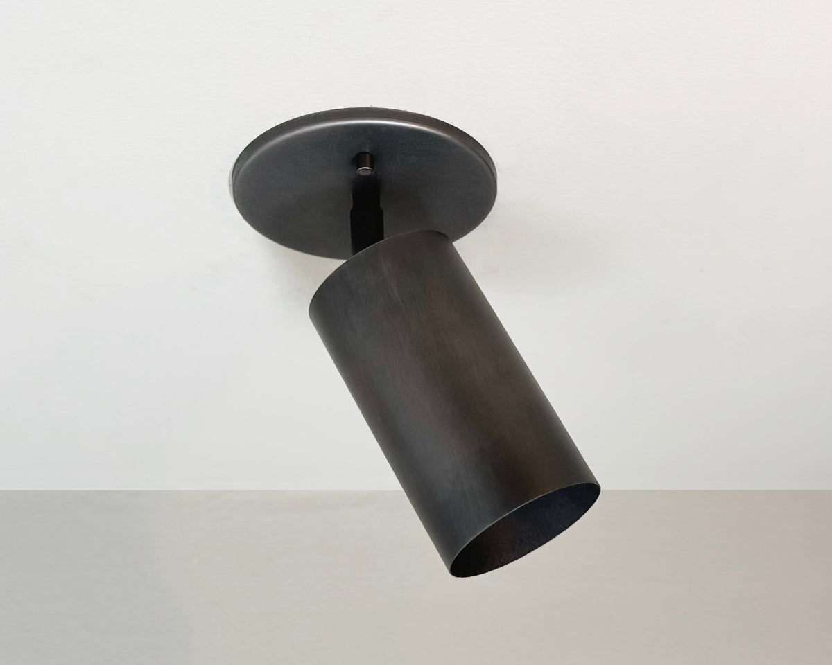 The Long Made Co. Cove Adjustable Spot is a solid steel, cylindrical black ceiling spotlight with a simple modern design, mounted on a white ceiling and angled downward—ideal for minimalist or vaulted lighting setups.