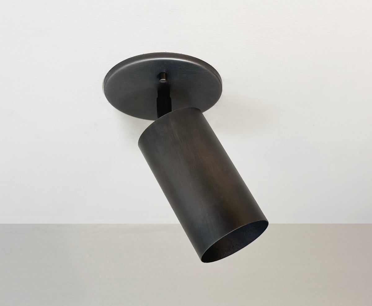 The Long Made Co. Cove Adjustable Spot, a black cylindrical ceiling spotlight from the Cove Series, is mounted on a round base against a white ceiling, angled down and to the side, showcasing modern minimalist elegance.