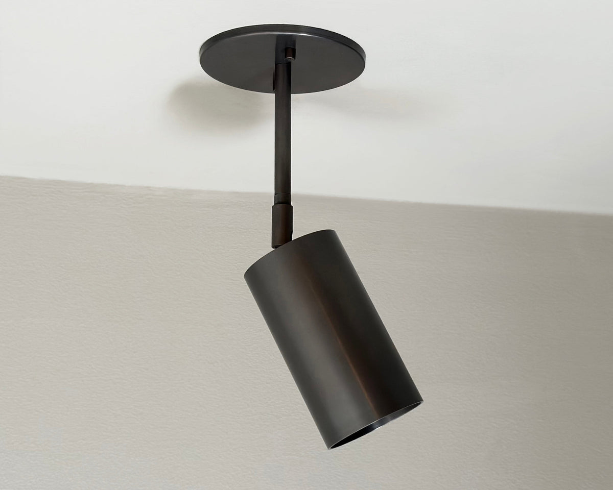 The Highland Drop Spot by Long Made Co. is a modern cylindrical black ceiling spotlight on a round base, mounted to a white ceiling and angled downward for focused task lighting.