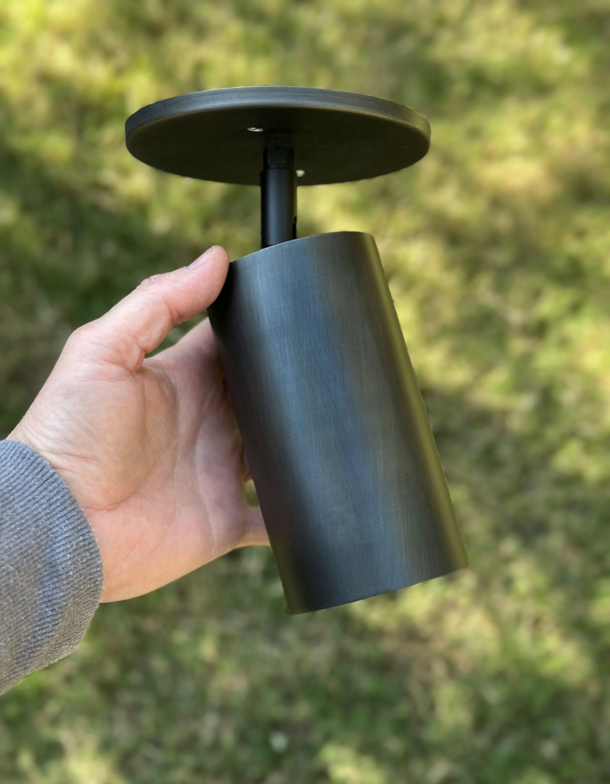 A hand holds the Long Made Co. Cove Adjustable Spot, a cylindrical black solid steel downlight with a round mounting plate, set against a blurred grassy background.