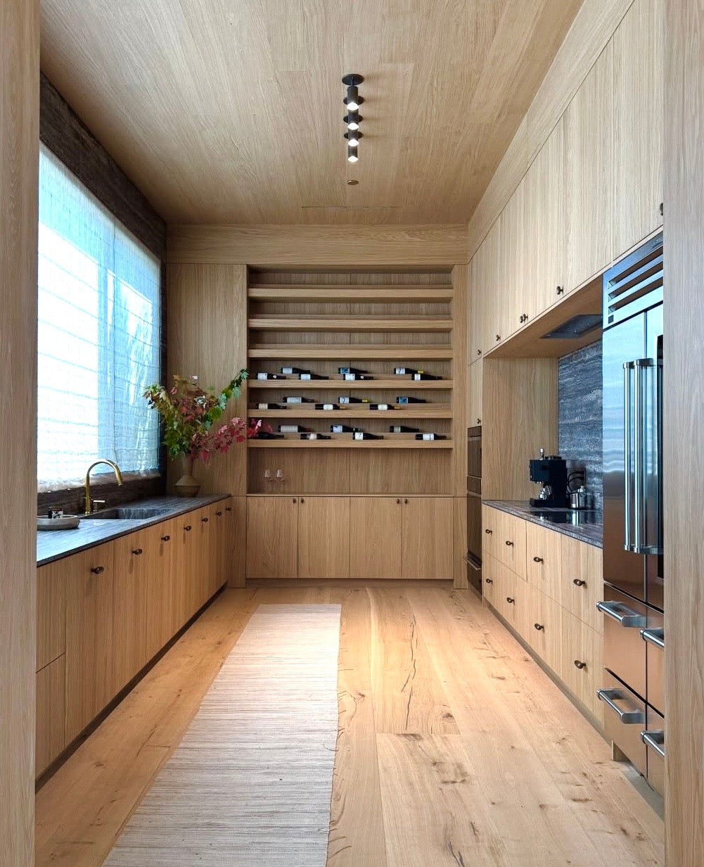 A modern kitchen with light wood cabinets, open shelves with jars, stainless steel appliances, and the Long Made Co. Brass Cylinder Adjustable Spot - Top Mount lighting. Sheer curtains, a beige runner, and flowers by the sink complete the look.