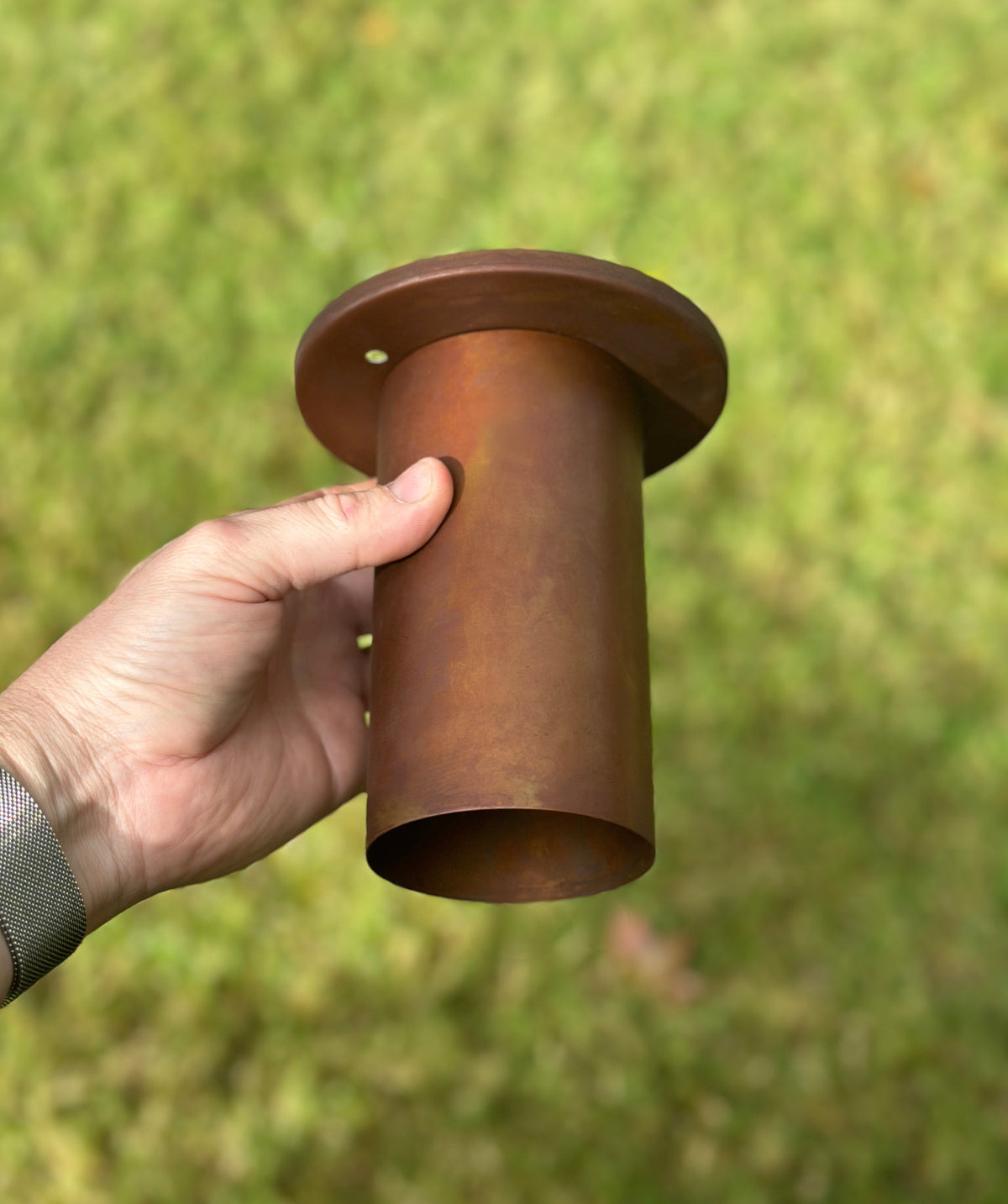 A hand holds the Long Made Co. Cove Flush Spot - Oxidized Rust, a cylindrical metal fixture with a flange, evoking minimalist lighting design, set against a blurred grassy background.