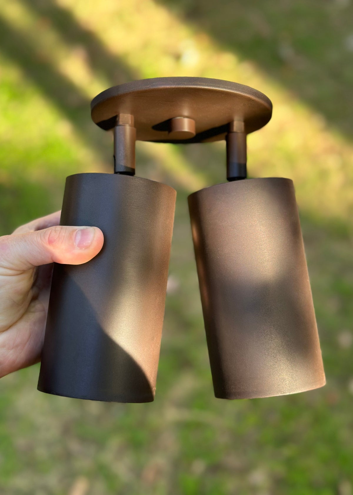 A hand holds the Long Made Co. Cove Double / Triple Spot, a bronze dual-cylinder outdoor light with a circular base and minimalist design, displayed outside on grass.