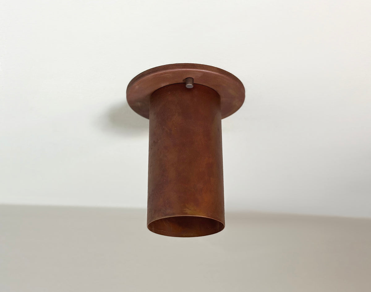 The Cove Flush Spot - Oxidized Rust by Long Made Co. is a cylindrical, industrial-style steel light fixture with a rust finish, mounted on a white ceiling and featuring an open end facing downward for minimalist lighting.