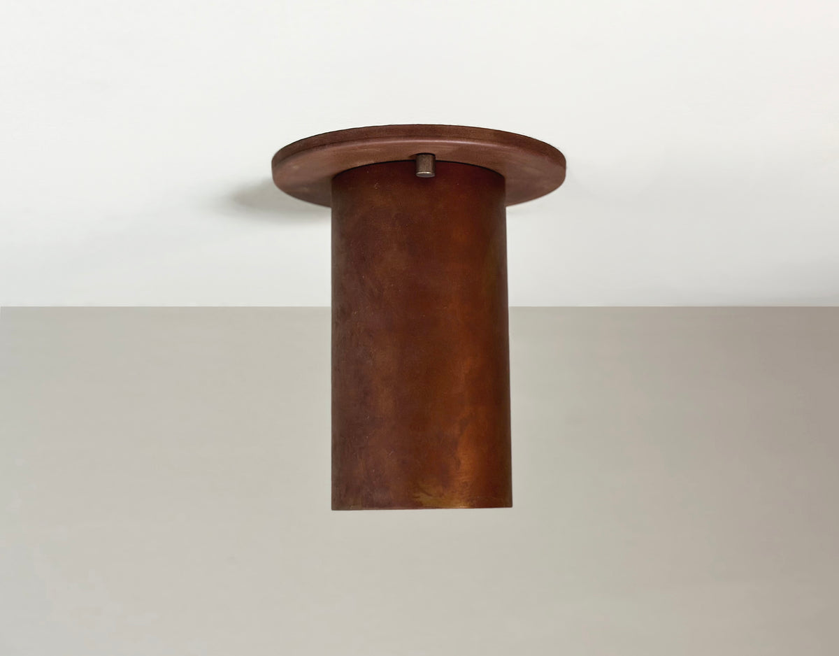 The Cove Flush Spot - Oxidized Rust by Long Made Co. is a cylindrical brown oxidized steel ceiling light with a flat round base, ideal for minimalist spaces, shown on a white ceiling against a light gray wall.