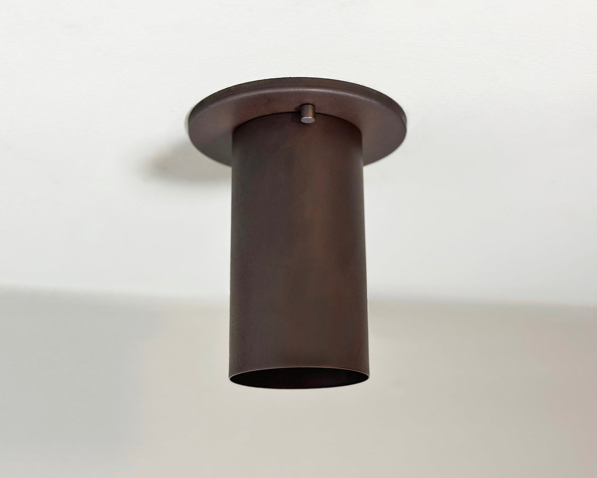 A cylindrical, dark brown Cove Flush Spot by Long Made Co. is mounted on a white ceiling. This solid steel downlight features an open bottom and a clean, modern design inspired by minimalist spotlights.
