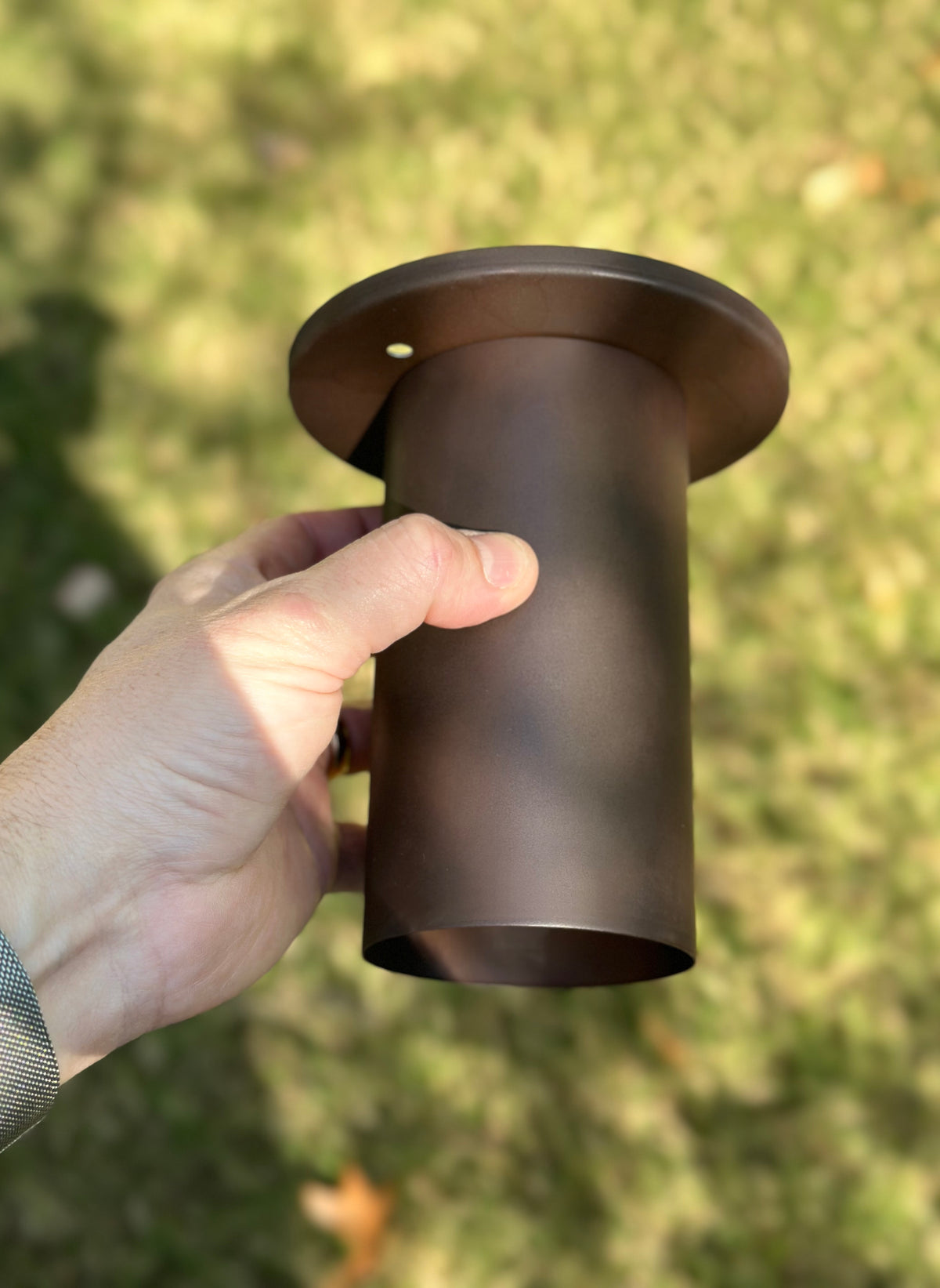 A hand holds the Cove Flush Spot by Long Made Co., a solid steel downlight with flange, against blurred grass in daylight—perfect for minimalist spotlights or precise task lighting.