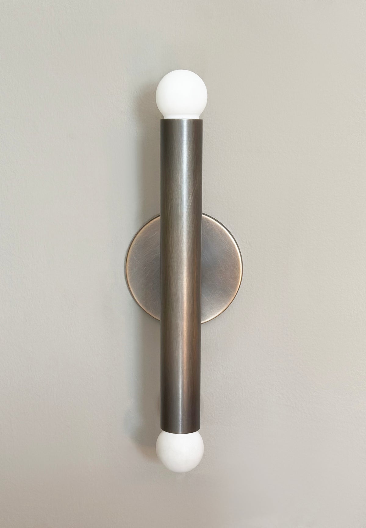 The Miro 2 - 12" Sconce by Long Made Co. features a brushed brass cylindrical body with white globe bulbs on each end, mounted vertically on a light beige wall—ideal for modern, stylish vanity lighting.