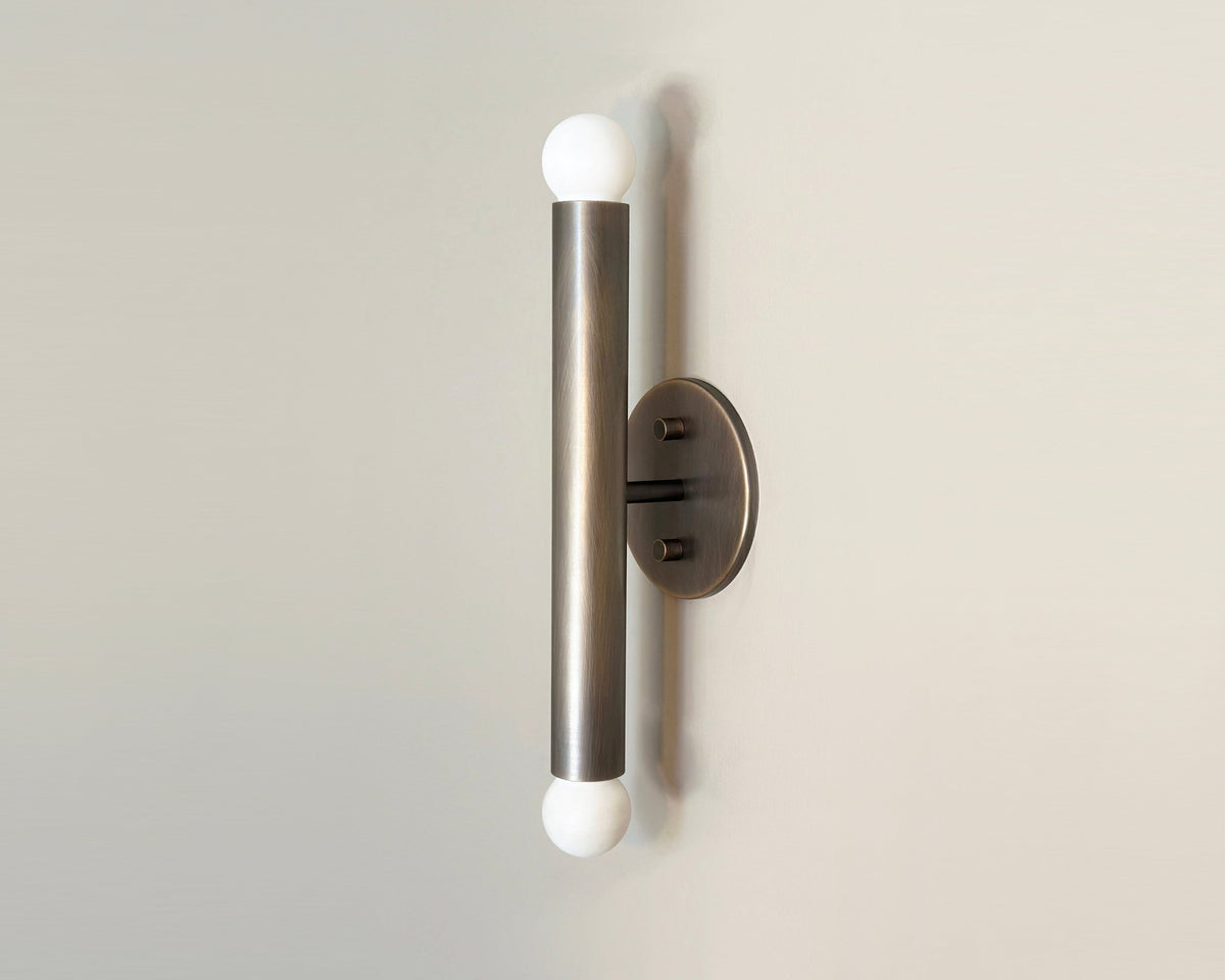 A sleek 12-inch wall sconce with brass connection points and a central cylindrical light fixture, featuring a warm oxidized finish.