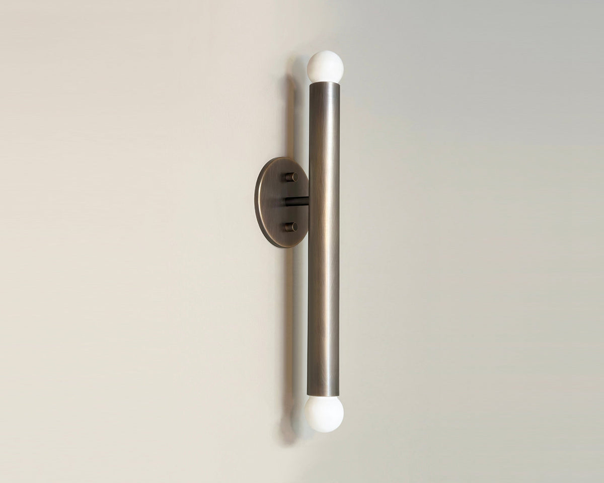 The Long Made Co. Miro 2 XL - 18" Sconce is a modern, cylindrical brass wall light with a brushed finish, mounted vertically and featuring round white bulbs on each end.