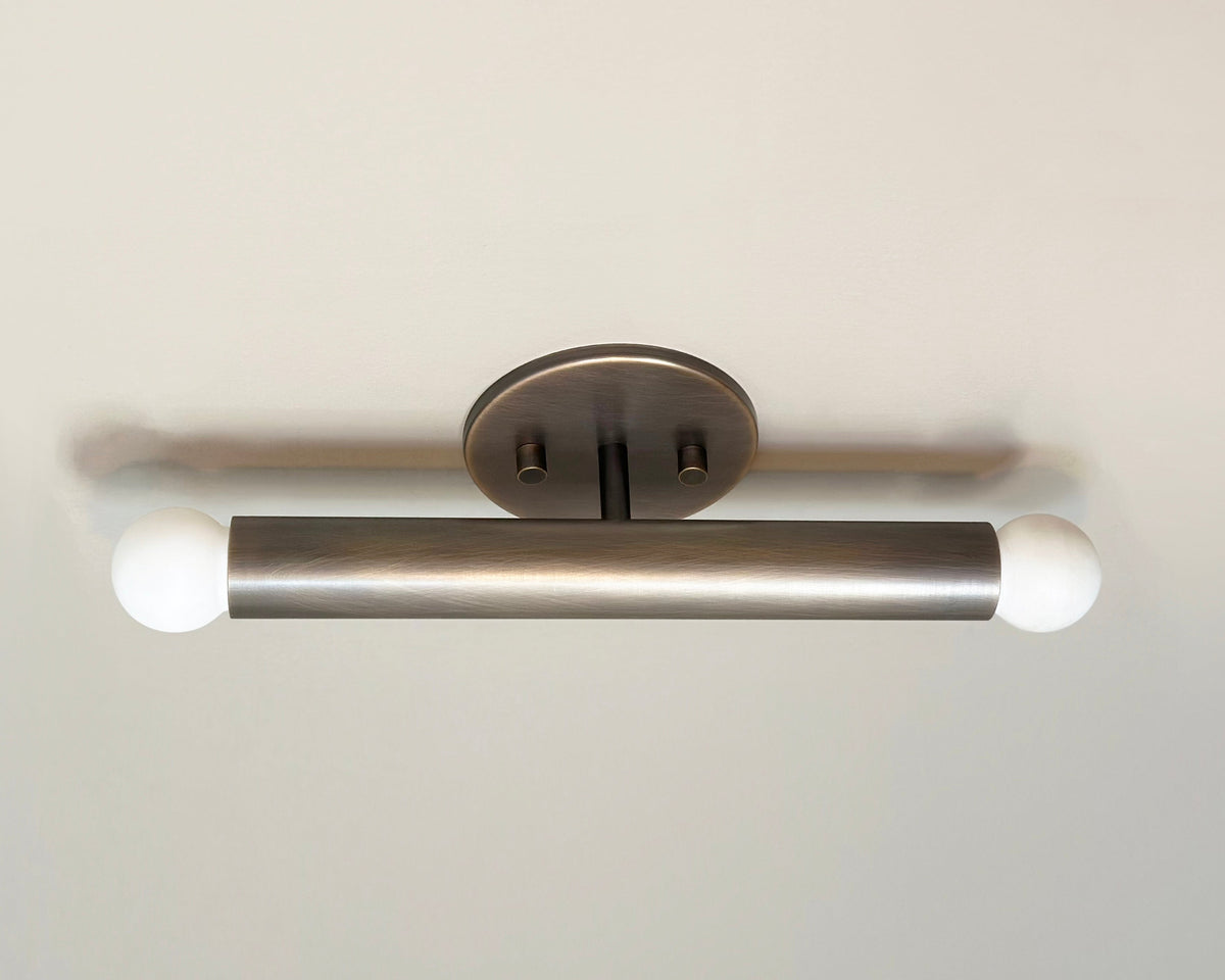 The Miro 2 - 12" Sconce by Long Made Co. is a modern, brushed metal cylindrical fixture with round white bulbs on each end, mounted horizontally on a circular base—perfect as a handmade vanity light for contemporary spaces.