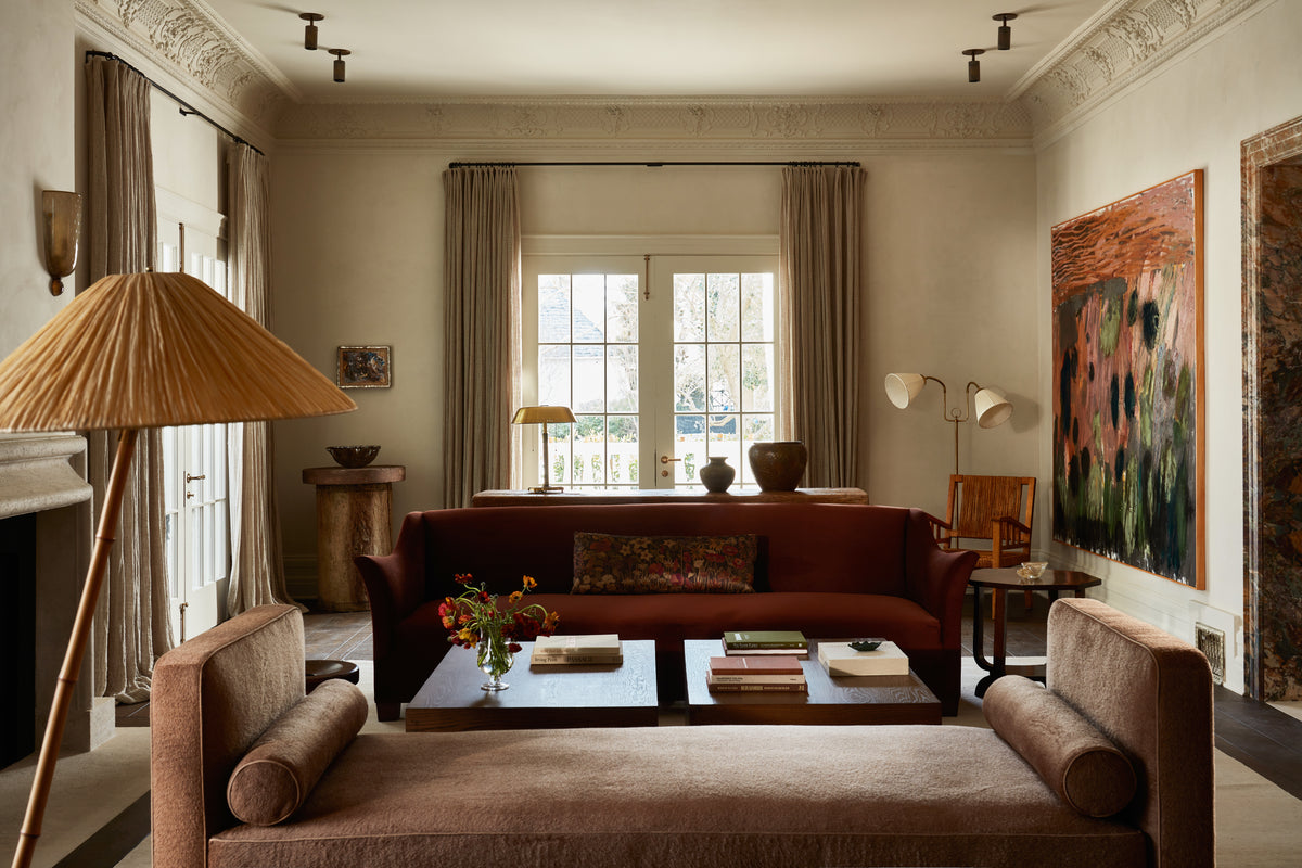 A warm living room with two brown sofas, a large coffee table, neutral curtains, wall art, and French doors. Mid-century modern lighting includes the Brass Cylinder Adjustable Spot - Top Mount by Long Made Co., adding elegance and cozy sophistication in earthy tones.