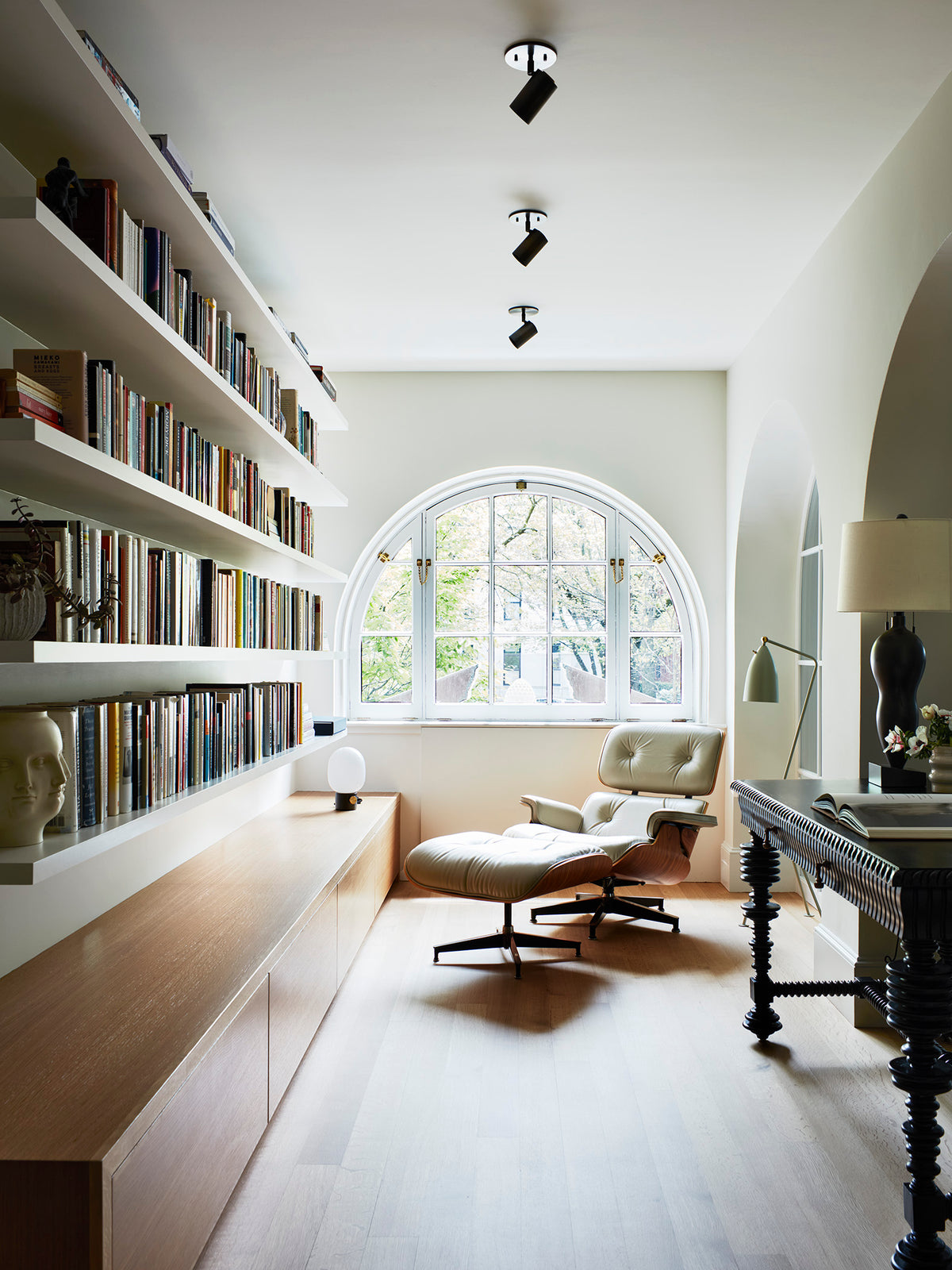 A bright, modern reading room with floor-to-ceiling bookshelves, wooden floors, a lounge chair and ottoman by an arched window, and a desk with a lamp. The space features Long Made Co.’s Brass Cylinder Adjustable Spot - Side Mount for mid-century lighting.