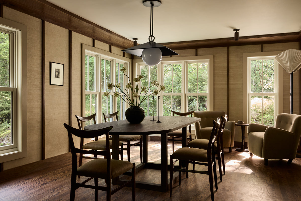 A dining room with large windows, wooden floors, a dark oval table, six chairs, an armchair, and a Long Made Co. Cove Adjustable Spot downlight. Greenery is visible outside and a vase with flowers sits on the table.