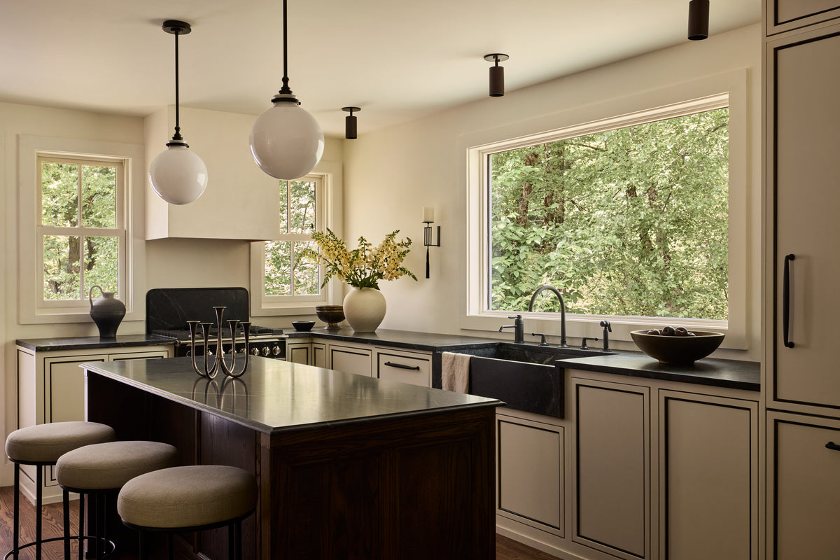 Modern kitchen featuring black countertops, cream cabinets, a dark wood island with three cushioned stools, Long Made Co. Cove Adjustable Spot lighting above, large windows framing green trees, and a sleek black sink under a big window.