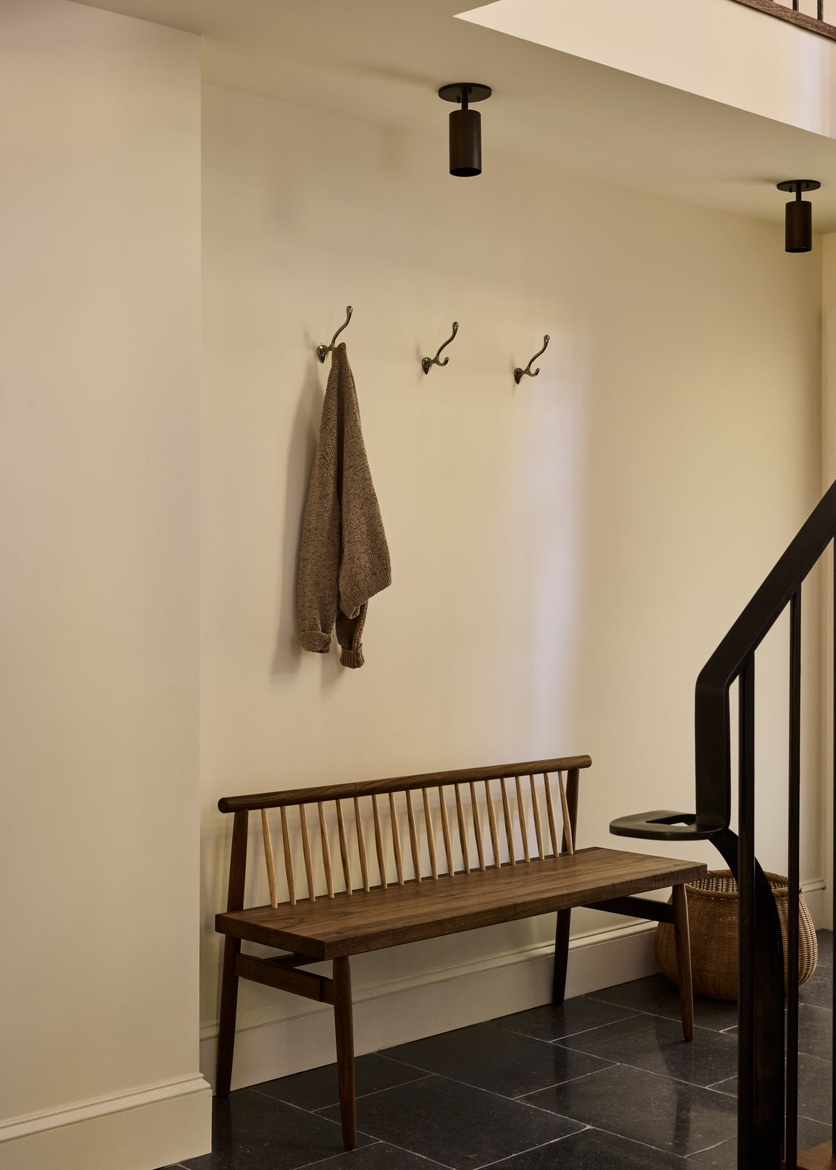 A minimalist hallway features a wooden bench, three wall hooks (one with a brown sweater), a woven basket, black tile flooring, and a staircase with a black railing, softly lit by the Highland Adjustable Spot from Long Made Co.