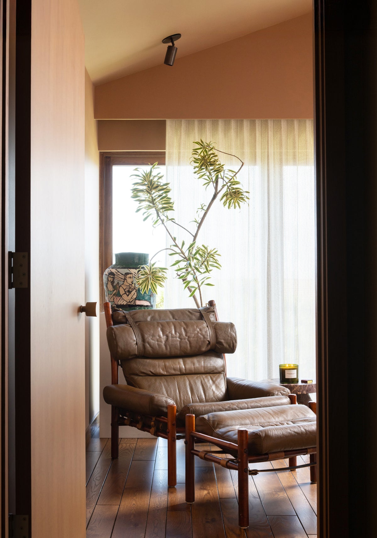 A cozy reading nook features a brown leather armchair and ottoman by a window with sheer curtains, leafy plant, small side table, and warm light from the Long Made Co. Brass Cylinder Adjustable Spot – Top Mount. A decorative vase and candle complete the space.