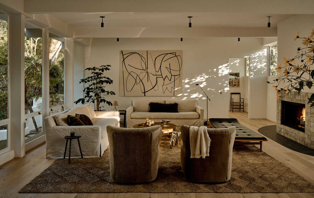 A cozy, modern living room with neutral tones, large windows, a fireplace, abstract art, armchairs, a sofa, and plants—illuminated overhead by the Brass Cylinder Drop Spot - Top Mount by Long Made Co., adding sleek mid-century flair.