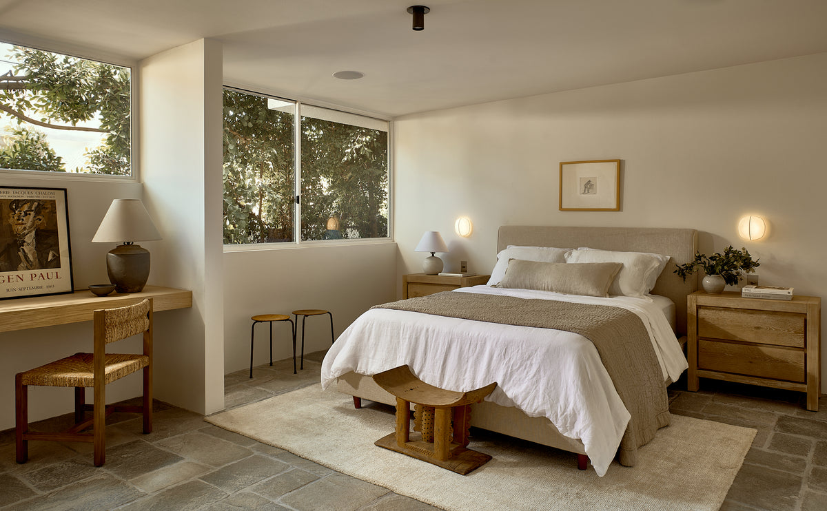 A cozy, modern bedroom features a large bed, wooden nightstands, minimal neutral decor, framed art, and ample natural light. Soft lighting from Long Made Co.'s Brass Cylinder Flush Spot adds a mid-century modern touch above the desk and chair.