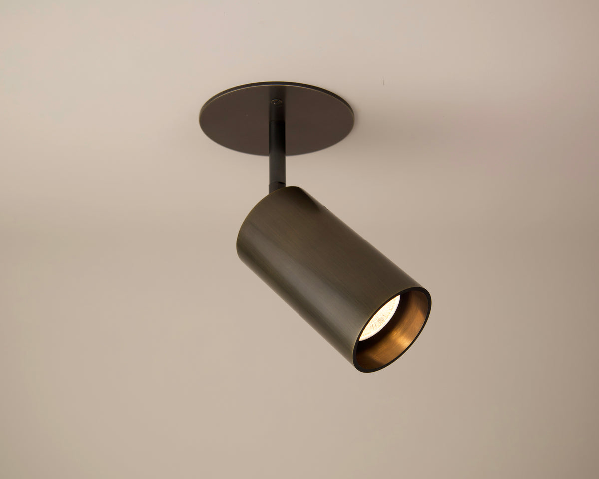 The Beau Laiton Drop Monopoint by Long Made Co. is a luxury cylindrical fixture, mounted on a plain light ceiling, angled downward and illuminated to cast a soft glow.