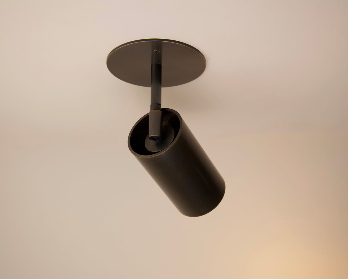 The Beau Laiton Drop Monopoint by Long Made Co. is a black cylindrical ceiling spotlight inspired by minimalist brass lighting, mounted on a white ceiling to cast focused downward light.