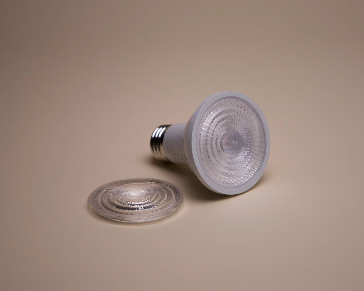 The Long Made Co. Bulb - Green Creative - PAR20 - E26 LED (2700K, 40/25° spread) stands upright beside its circular lens cover on a beige surface, highlighting its energy efficiency and warm glow.