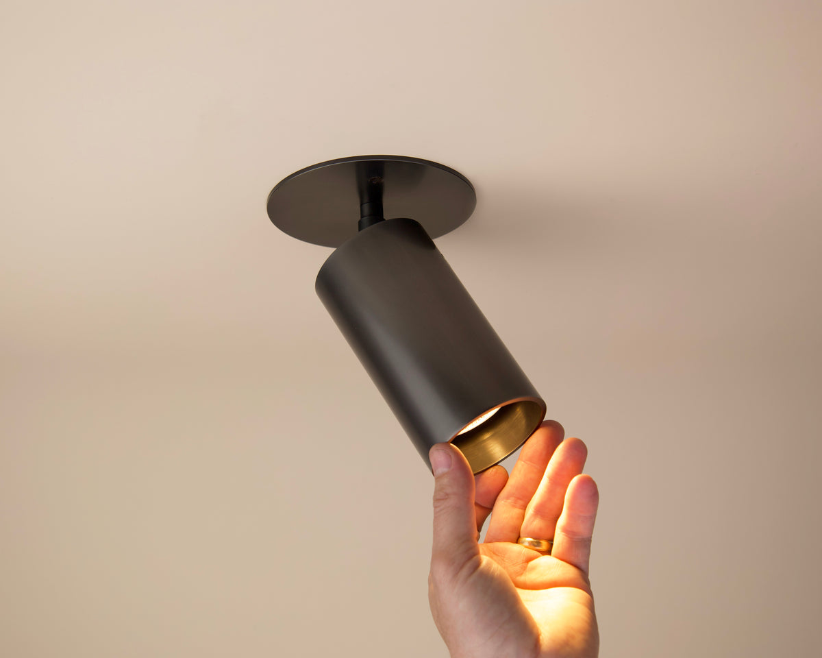 A hand reaches up to adjust the Long Made Co. Beau Laiton Monopoint, a sleek cylindrical black luxury flush mount ceiling light fixture set against a beige ceiling.