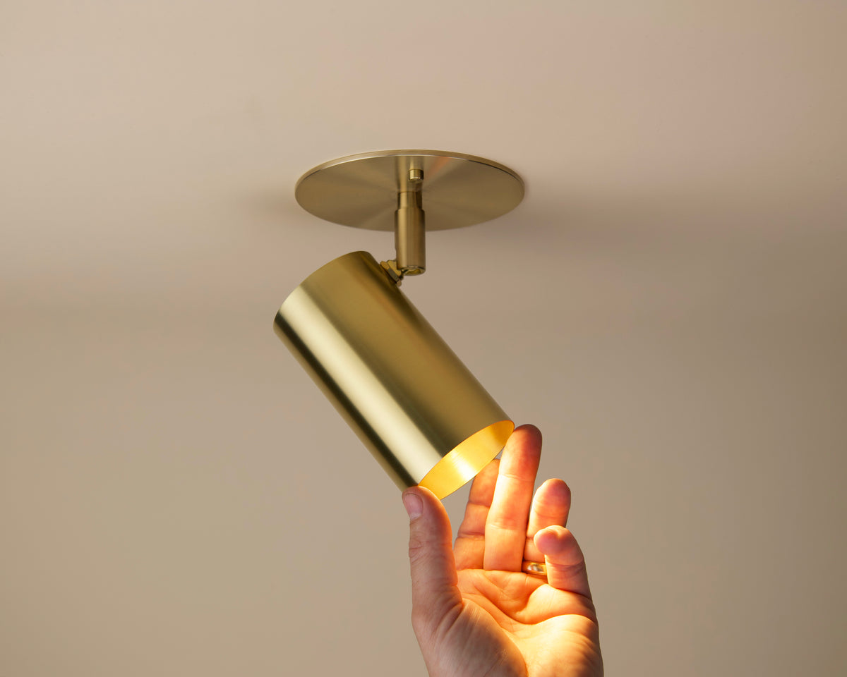 A hand adjusts the Long Made Co. Brass Cylinder Adjustable Spot - Side Mount, a modern brass ceiling downlight emitting warm light from a beige ceiling—the perfect mid-century modern lighting accent.