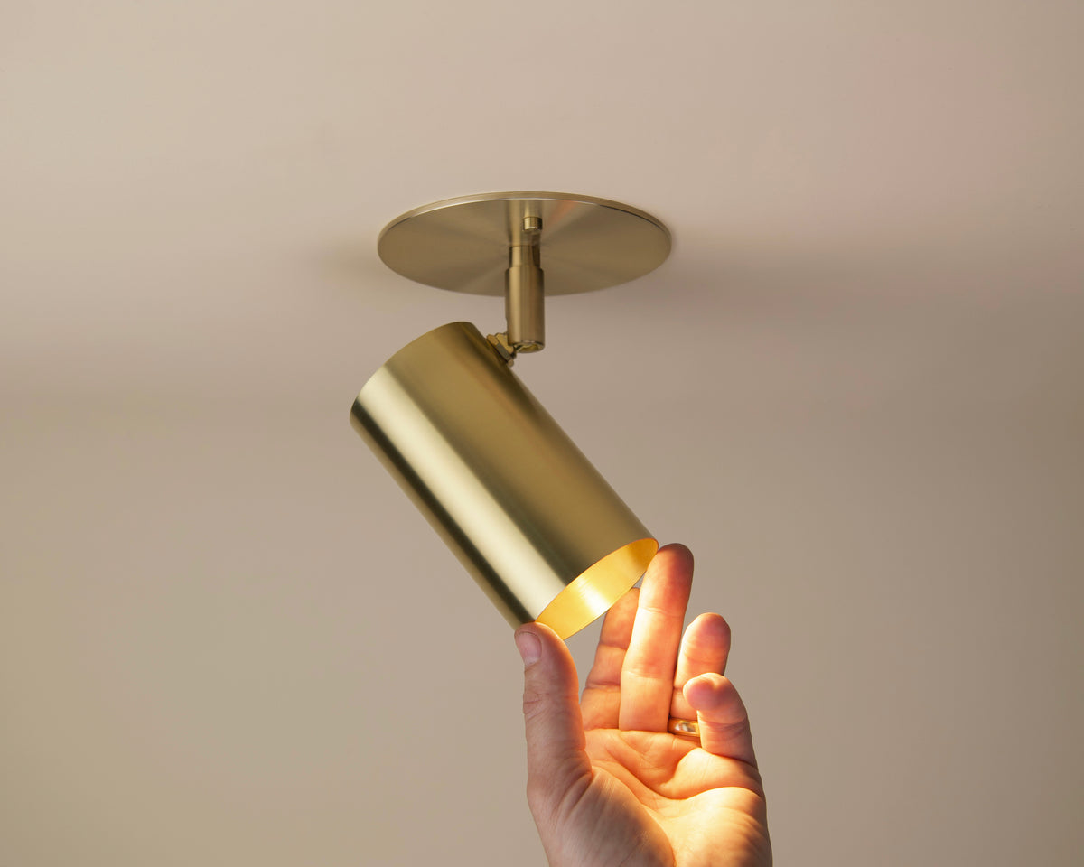 A hand adjusts the Long Made Co. Brass Cylinder Adjustable Spot - Side Mount fixture, mounted on a neutral-colored ceiling, embodying the sleek elegance of mid-century modern lighting.