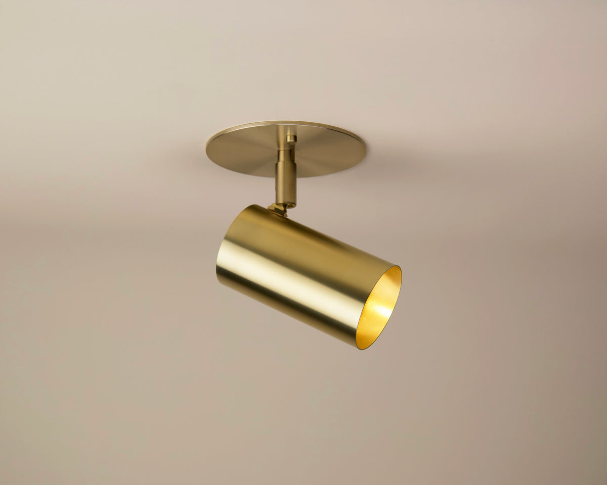The Brass Cylinder Adjustable Spot - Side Mount by Long Made Co. features a modern cylindrical brass shade on a round base, angled downward—ideal for mid-century modern lighting in any space.