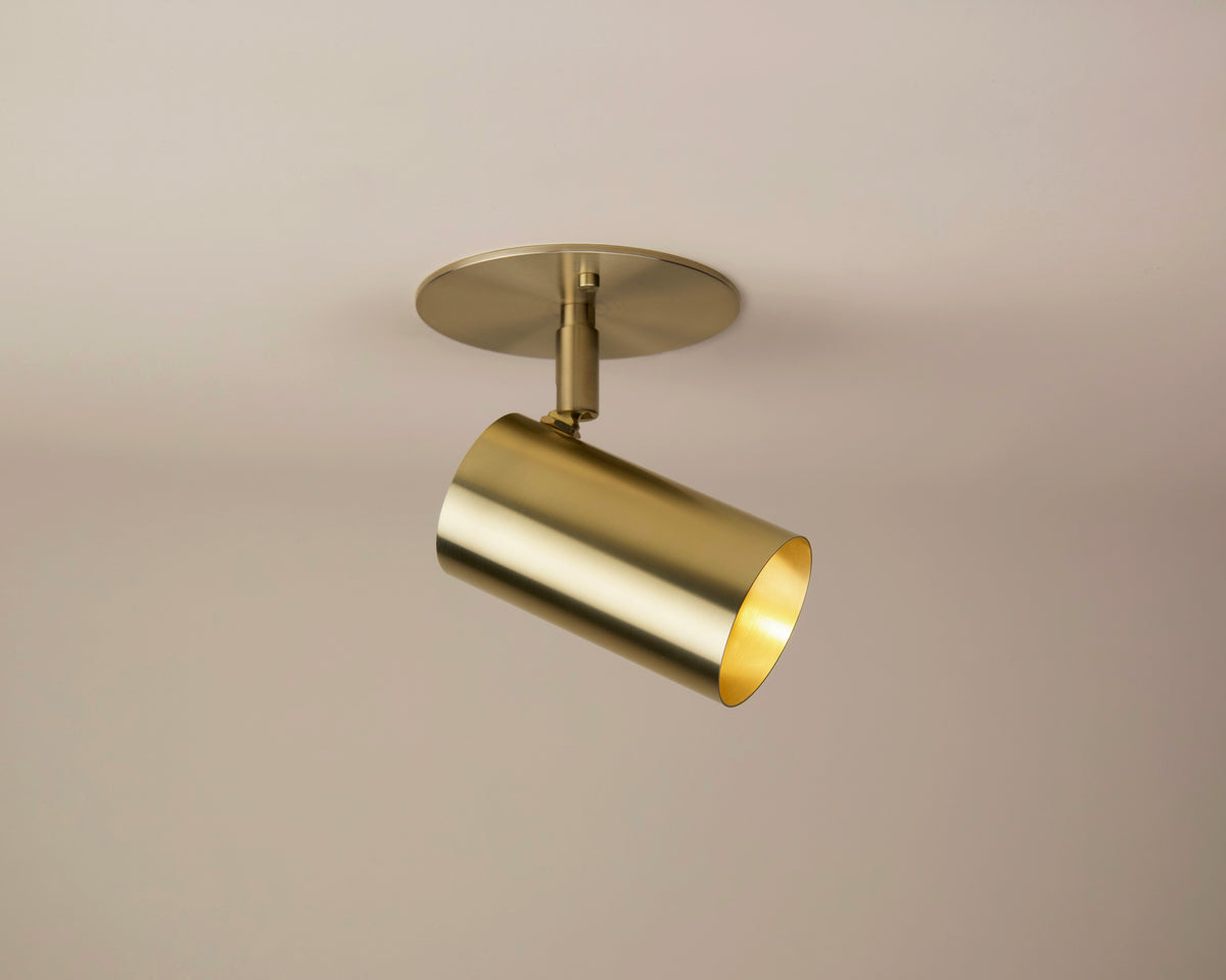 Mounted on a light-colored ceiling, the Brass Cylinder Adjustable Spot - Side Mount by Long Made Co. features a solid brass cylindrical form with a round base, casting a warm glow. This directional monopoint fixture combines functional adjustability with sleek style, making it an elegant choice for mid-century modern lighting schemes.