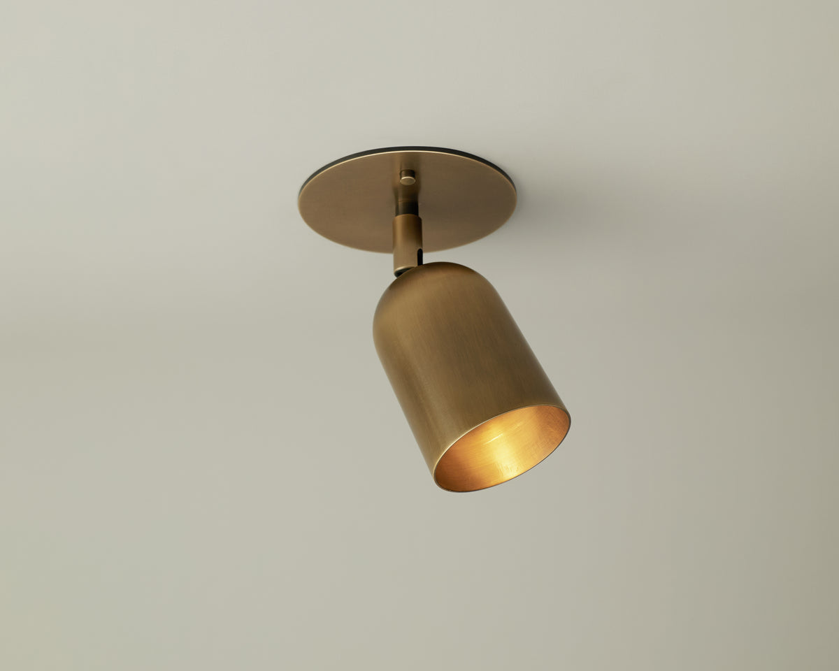 The Campana Adjustable Spot by Long Made Co. features a solid brass cylindrical shade on a round base, casting light against a plain ceiling—a timeless example of mid-century lighting.