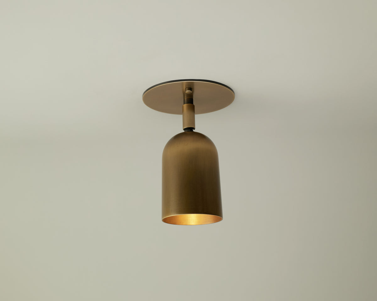 The Long Made Co. Campana Adjustable Spot, a brass-colored cup-shaped ceiling downlight, is mounted on a plain white ceiling and casts a warm glow downward.