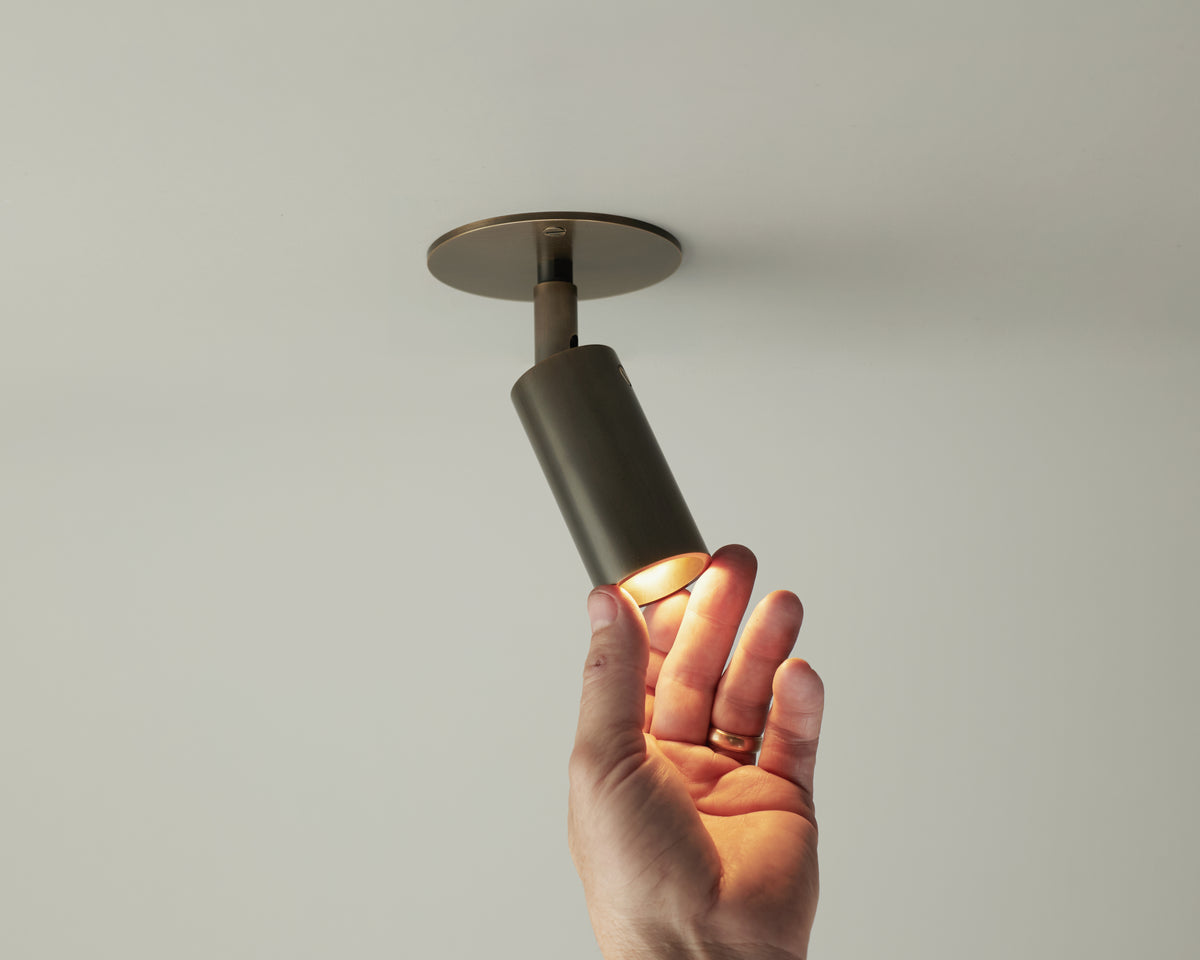 A hand adjusts the Micro Adjustable Spot by Long Made Co., its minimalist brass fixture casting a warm, focused light downward onto a plain white ceiling.